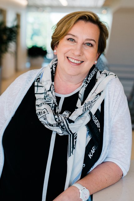 Photo of Kasia Hein-Peters: a professional Caucasian woman wearing a black top, white sweater, and black and white scarf. She is standing with her left elbow on a table.